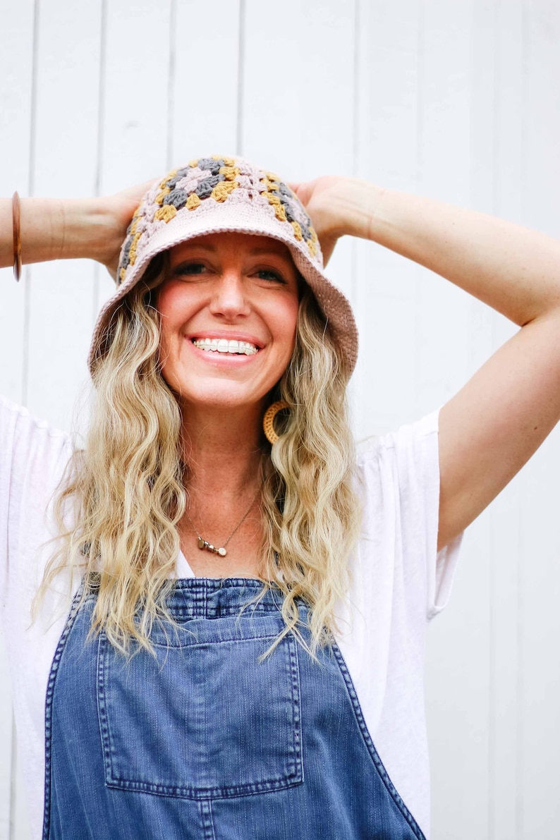 A blonde woman smiling wearing a bucket hat, a white t-shirt, and denim overalls with her hands-on the hat.