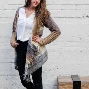 Puede incluir: Un cárdigan largo de crochet abierto en tonos marrón, beige y gris. El cárdigan presenta un patrón de rayas y flecos en el borde inferior. Las mangas son largas y ajustadas, y el estilo general es informal y cómodo.