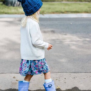 A blonde little girl standing on a puddle of water wearing a royal blue beginner child beanie, a white cardigan, a floral dress, and a pair of blue rain boots.