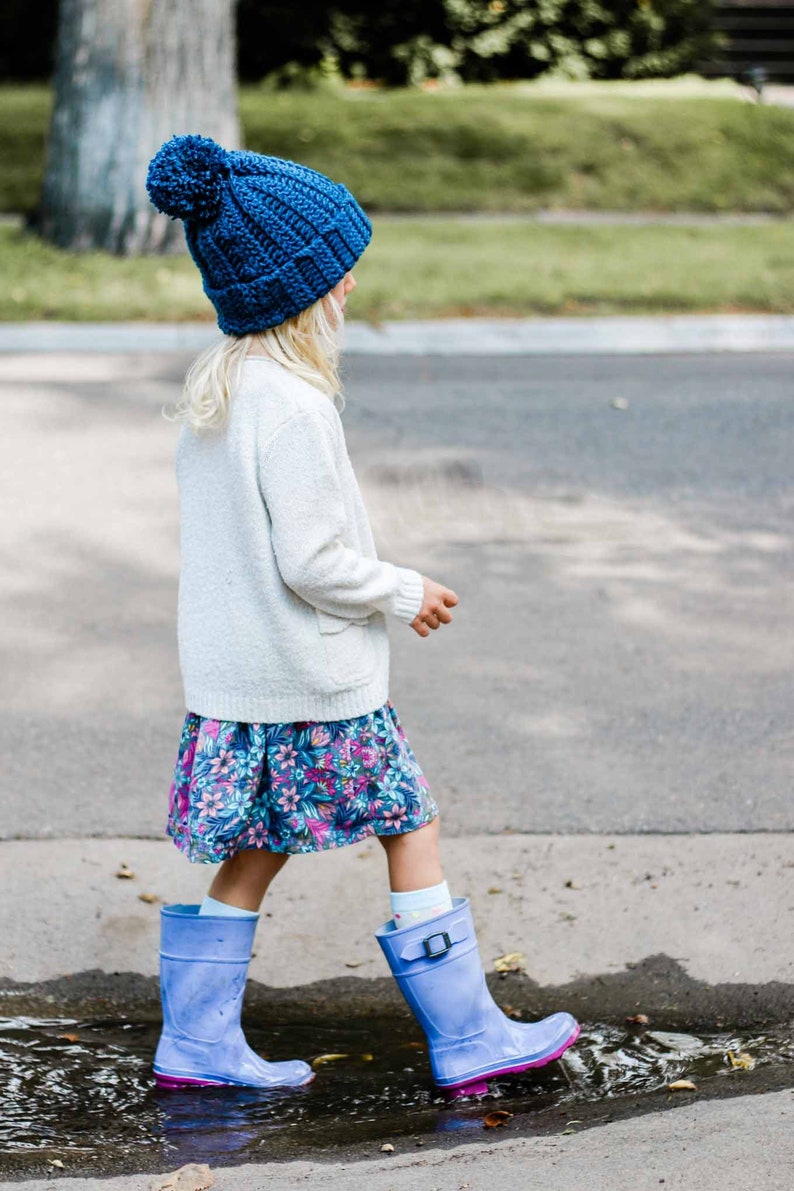 A blonde little girl standing on a puddle of water wearing a royal blue beginner child beanie, a white cardigan, a floral dress, and a pair of blue rain boots.
