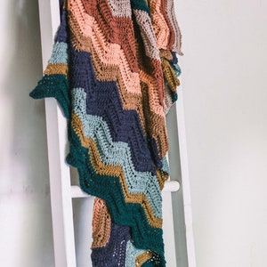 May include: A multi-colored crocheted blanket with a chevron pattern in shades of teal, brown, pink, and gray. The blanket is draped over a white wooden ladder.