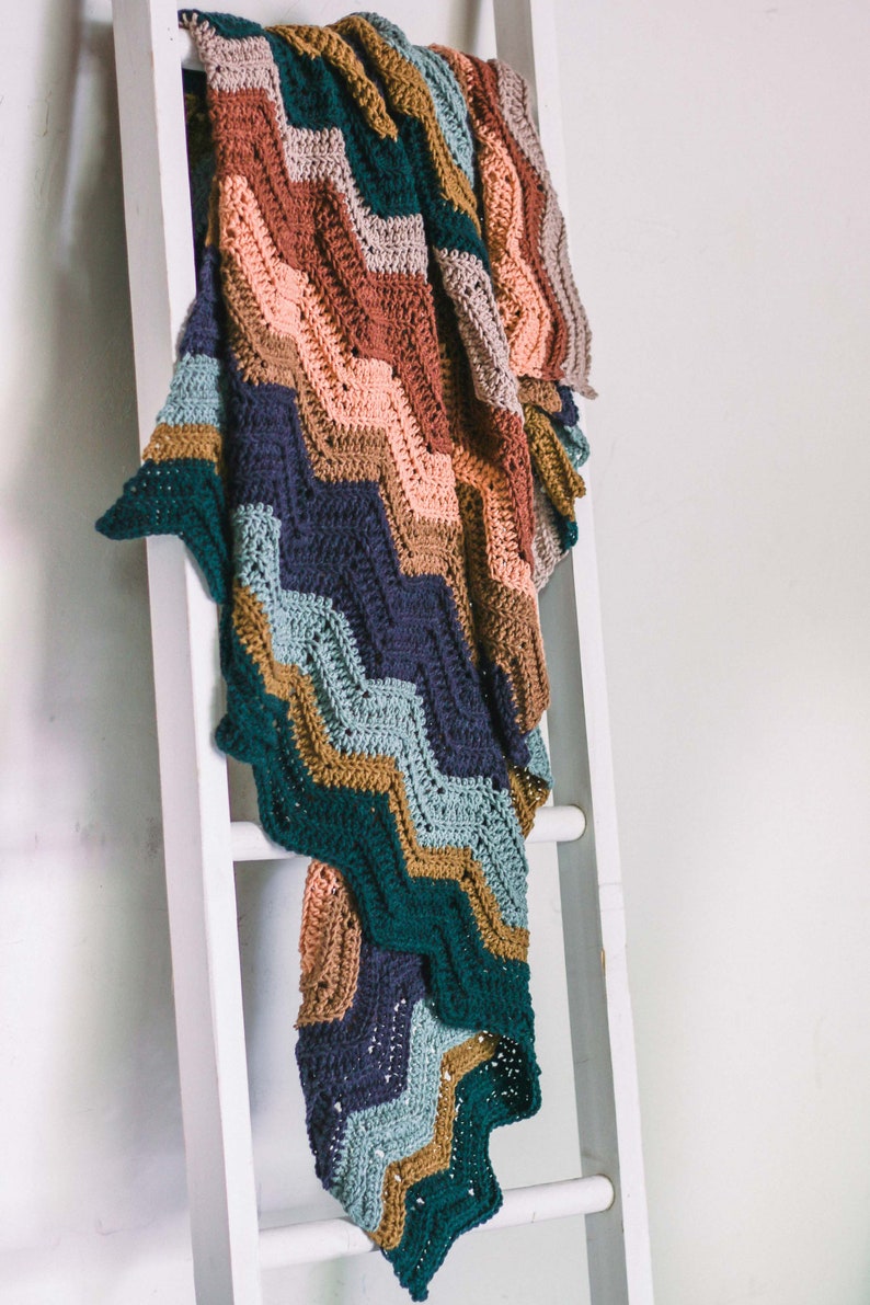 May include: A multi-colored crocheted blanket with a chevron pattern in shades of teal, brown, pink, and gray. The blanket is draped over a white wooden ladder.