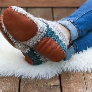 A woman with her legs crossed sitting on a fur rug wearing denim jeans and a pair of white beginner crochet slippers with leather soles.
