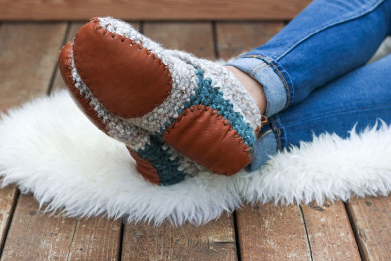 A woman with her legs crossed sitting on a fur rug wearing denim jeans and a pair of white beginner crochet slippers with leather soles.