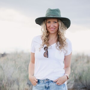 A blonde woman is smiling, standing on a field wearing a green crochet sun hat, a white t-shirt with sunglasses at the collar, and denim jeans with her hands on the front pockets.