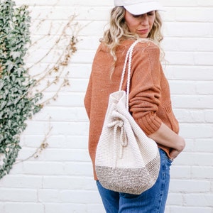 A blonde woman standing with her back slightly to the camera, smiling, wearing a cap, an orange sweater, denim jeans, and a crochet drawstring purse on her shoulders.