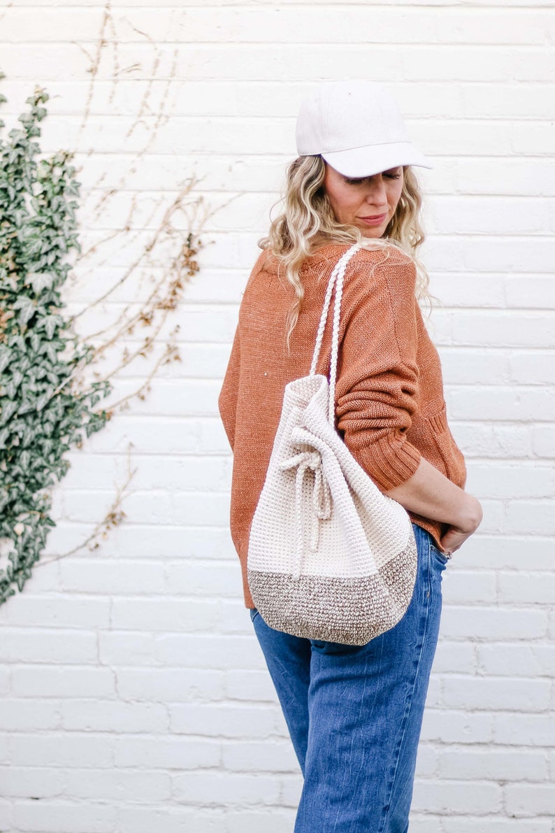 A blonde woman standing with her back slightly to the camera, smiling, wearing a cap, an orange sweater, denim jeans, and a crochet drawstring purse on her shoulders.