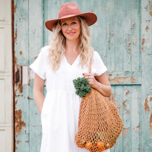 A blonde woman smiling, standing in front of a green door wearing a fedora hat and a white dress carrying a cotton crochet bag with  fruits and vegetables on her shoulder.