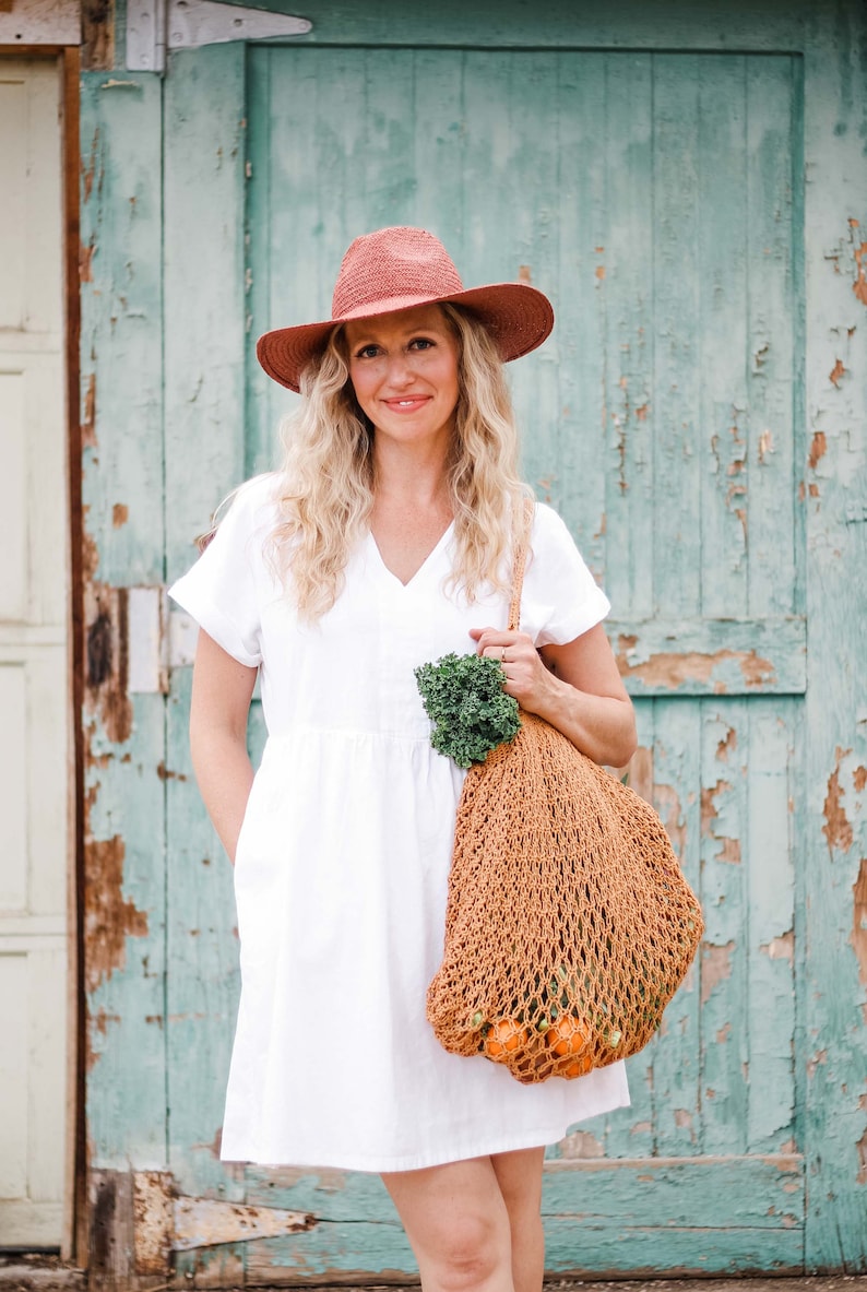 A blonde woman smiling, standing in front of a green door wearing a fedora hat and a white dress carrying a cotton crochet bag with  fruits and vegetables on her shoulder.
