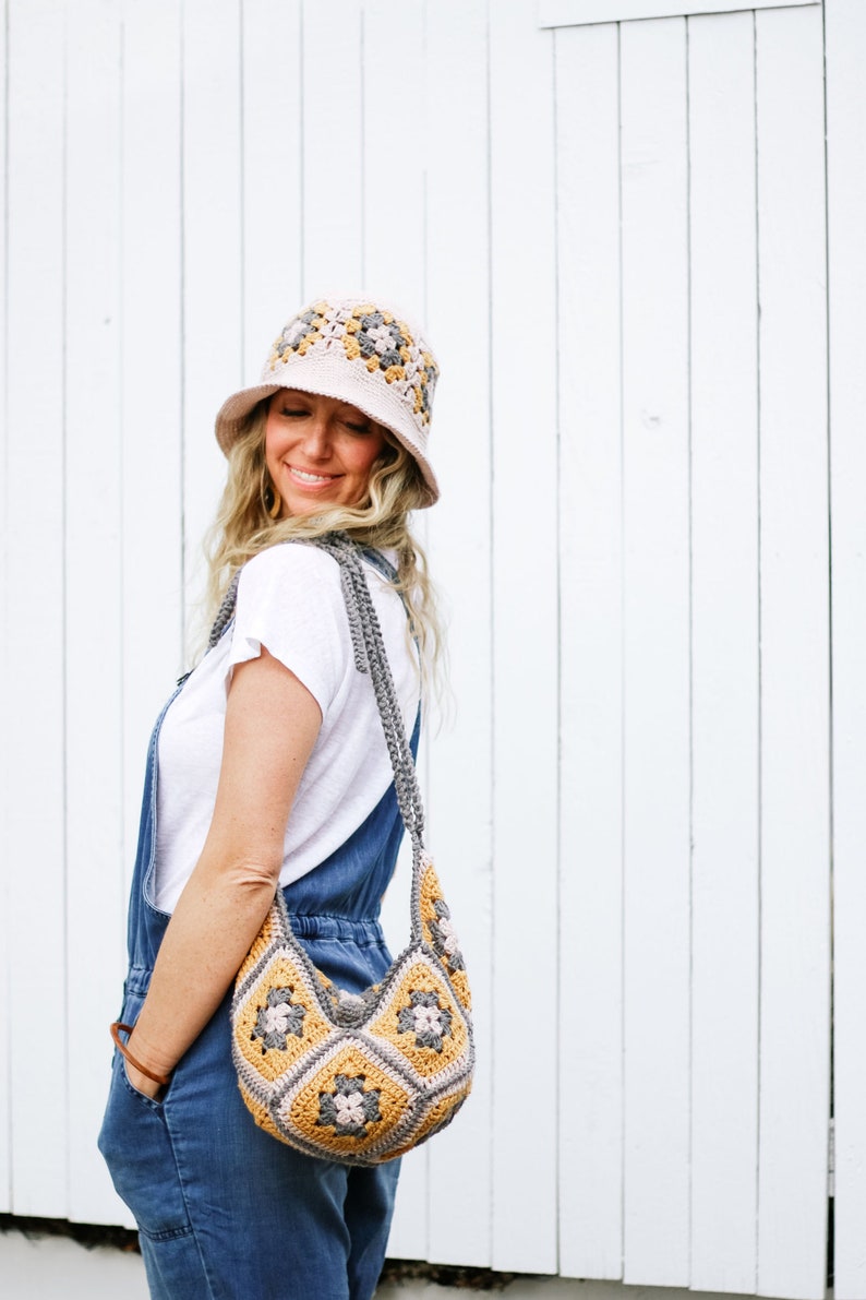 A blonde woman stands in front of a white wall, looking down and smiling. She is wearing a crochet sun hat with a matching bag on her shoulder, a white T-shirt, and a denim jumper with her hand on the front pocket.
