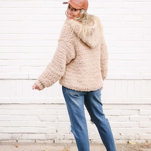 A blonde woman smiles, stands with her back to the camera, and wears a brown cap, a beige oversized sweater with a hood, denim jeans, and a pair of white sneakers.
