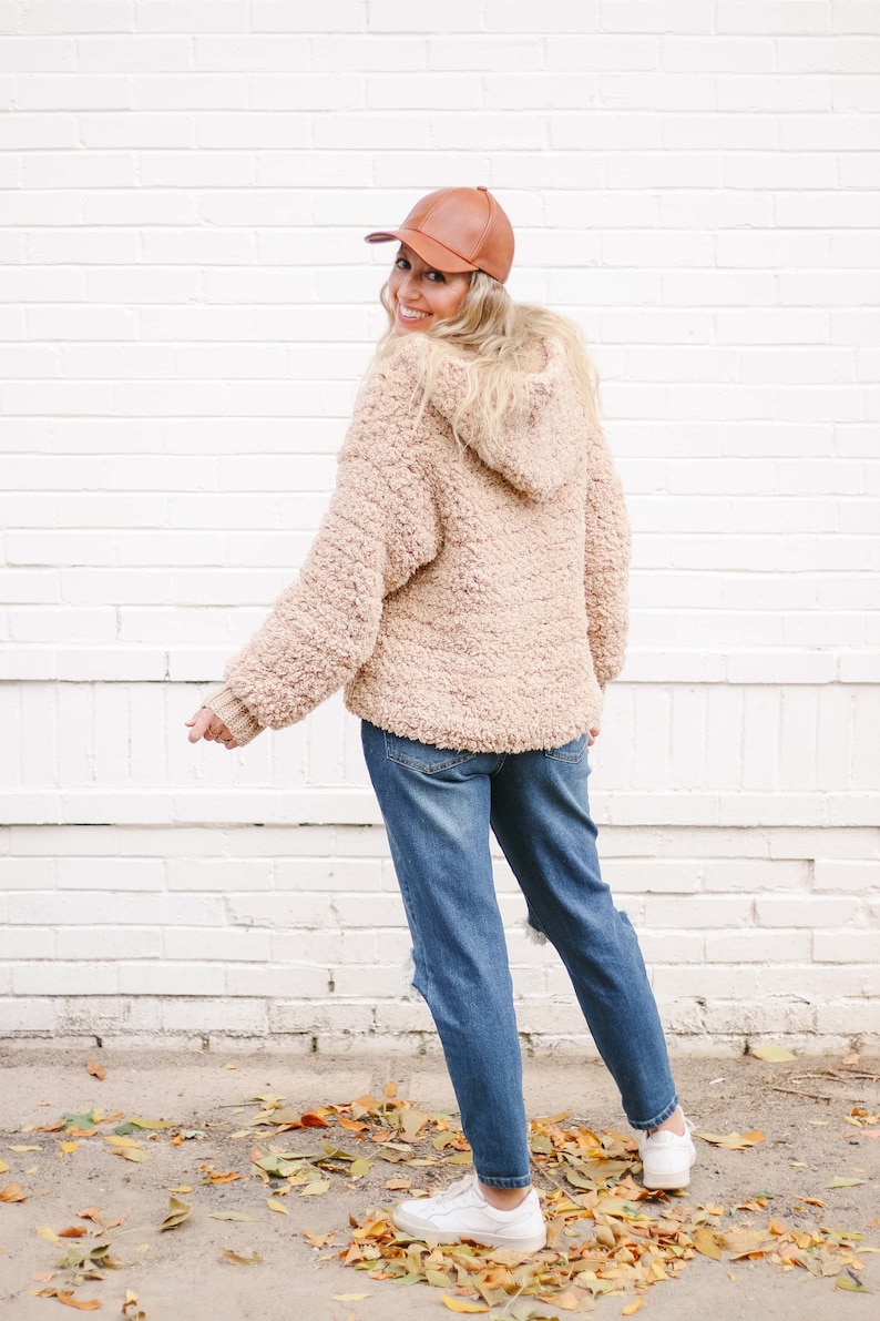 A blonde woman smiles, stands with her back to the camera, and wears a brown cap, a beige oversized sweater with a hood, denim jeans, and a pair of white sneakers.