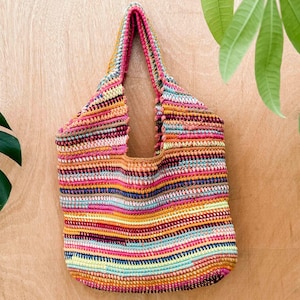 Könnte beinhalten: Eine farbenfrohe, handgehäkelte Tragetasche mit einem gestreiften Muster in Rosa, Orange, Gelb, Blau und Braun. Die Tasche hat einen stabilen Griff und wird vor einem hellbraunen Holzgrund präsentiert.