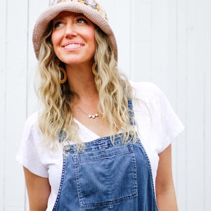 A blonde woman is smiling, looking up, wearing a hat with a brim, a white shirt, a necklace, and a denim jumper.