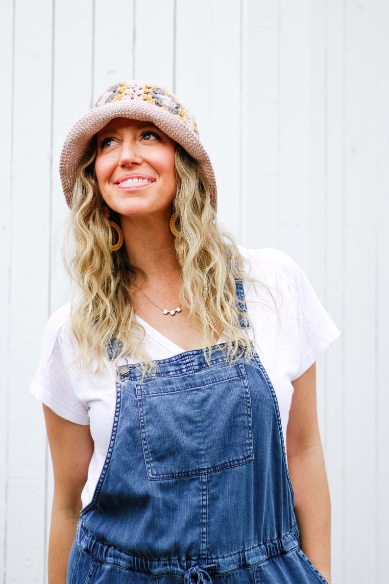 A blonde woman is smiling, looking up, wearing a hat with a brim, a white shirt, a necklace, and a denim jumper.