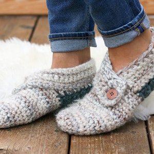 A woman standing on a fur rug wearing denim jeans and a pair of beginner crochet slippers with leather soles and a button detail with one foot on tiptoe.