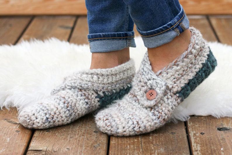 A woman standing on a fur rug wearing denim jeans and a pair of beginner crochet slippers with leather soles and a button detail with one foot on tiptoe.