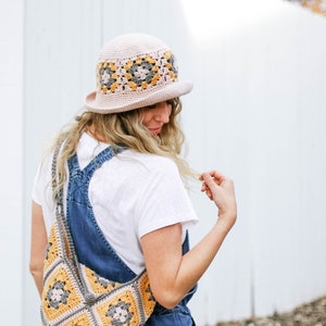 A blonde woman is standing with her back to the camera, looking down and smiling while wearing a granny square hat with a matching bag, a white t-shirt, and denim overalls.