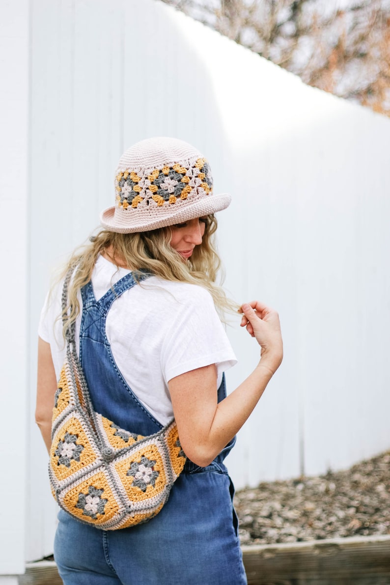 A blonde woman is standing with her back to the camera, looking down and smiling while wearing a granny square hat with a matching bag, a white t-shirt, and denim overalls.