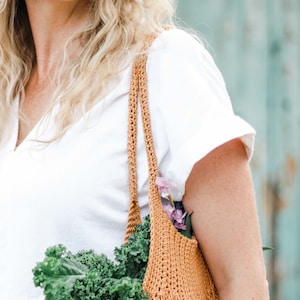 A blonde woman carrying a mesh market bag full of vegetables and flowers.