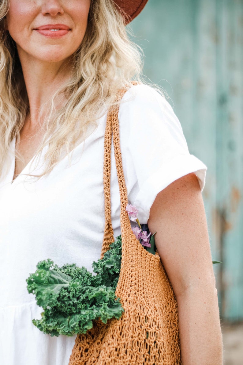 A blonde woman carrying a mesh market bag full of vegetables and flowers.