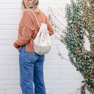 A blonde woman standing with her back slightly to the camera, smiling, wearing a cap, an orange sweater, denim jeans, and a mini backpack on her shoulders.