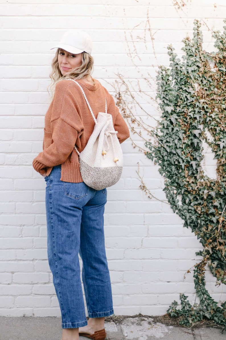 A blonde woman standing with her back slightly to the camera, smiling, wearing a cap, an orange sweater, denim jeans, and a mini backpack on her shoulders.