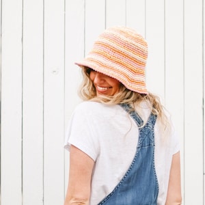 A blonde woman smiles with her back to the camera, wearing an orange-themed packable sun hat, denim overalls, and a white T-shirt.