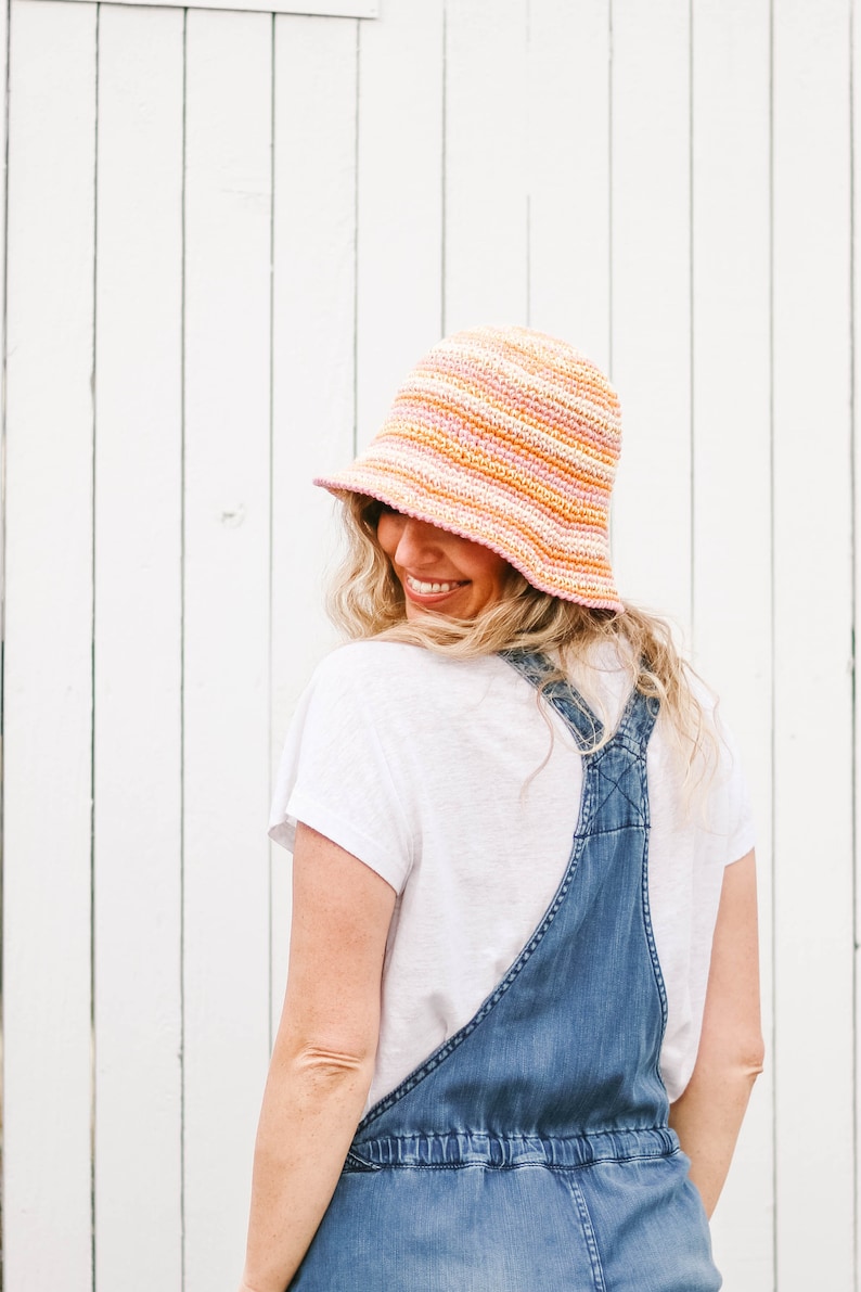 A blonde woman smiles with her back to the camera, wearing an orange-themed packable sun hat, denim overalls, and a white T-shirt.