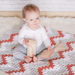 May include: A baby sitting on a gray and orange crocheted blanket, playing with wooden blocks. The baby is wearing a white shirt and gray pants.