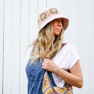 A blonde woman standing, looking sideways wearing a granny square hat with a matching bag, a white top, and a denim jumper, with her hand on the leather strap of the bag on her shoulder.