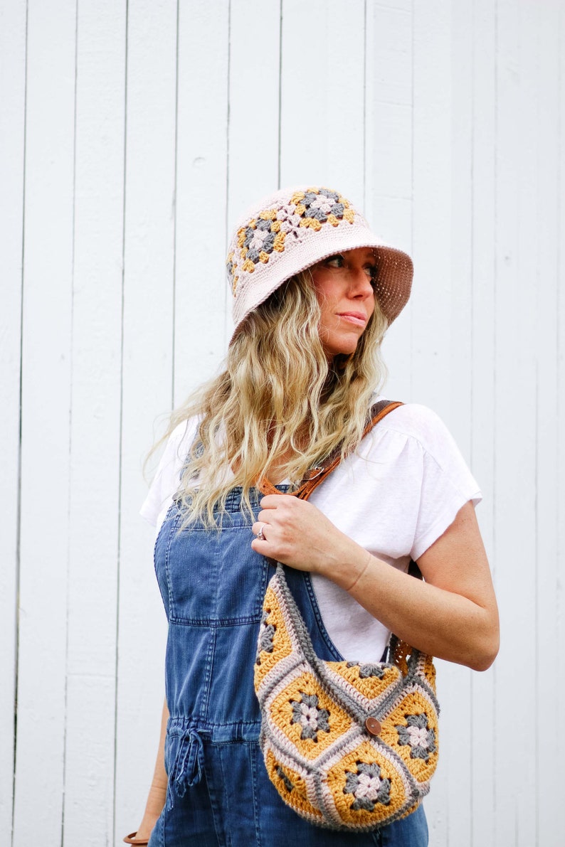 A blonde woman standing, looking sideways wearing a granny square hat with a matching bag, a white top, and a denim jumper, with her hand on the leather strap of the bag on her shoulder.