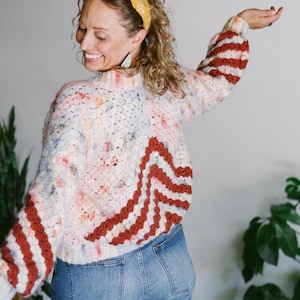 May include: A woman wearing a white and red crocheted sweater with a yellow headband. The sweater has a geometric pattern and the sleeves are striped.