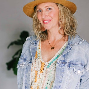 A blonde woman looking smiling at the camera. She is wearing a v-neck crochet tee, jean jacket, and tan hat.