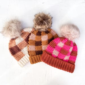 May include: Three handmade crochet beanies with faux fur pom-poms. The beanies feature a checkered pattern in shades of brown, pink, and cream. The pom-poms are in matching neutral colors. The hats are displayed on a white surface.