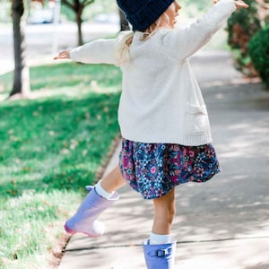 A blonde little girl dancing is wearing a blue beginner child beanie, a white cardigan, a floral dress, and blue rain boots.