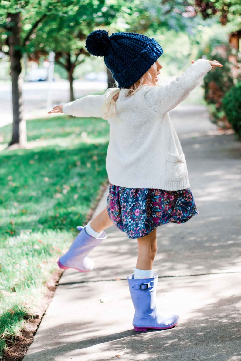 A blonde little girl dancing is wearing a blue beginner child beanie, a white cardigan, a floral dress, and blue rain boots.