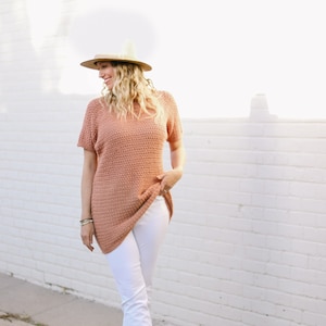 May include: A woman wearing a peach-coloured crochet top with white trousers and brown leather shoes. She is wearing a wide-brimmed straw hat.