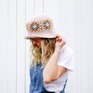 A blonde woman standing in front of a white wall wearing crochet sun hat, a white t-shirt, and denim overalls with her hand on the brim of the hat.