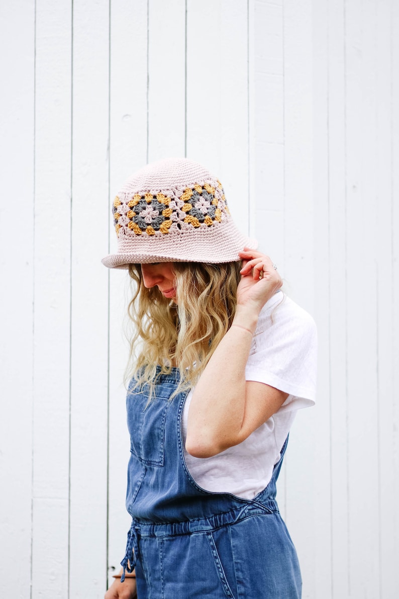 A blonde woman standing in front of a white wall wearing crochet sun hat, a white t-shirt, and denim overalls with her hand on the brim of the hat.