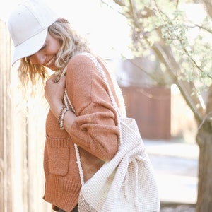 A blonde woman smiling, carrying a crochet backpack on her shoulder. She is wearing a white cap, an orange sweater and denim jeans.