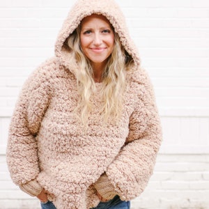 A blonde woman wearing a sherpa hoodie with the hood on her head, and denim jeans with her hands on the front pockets of her jeans.
