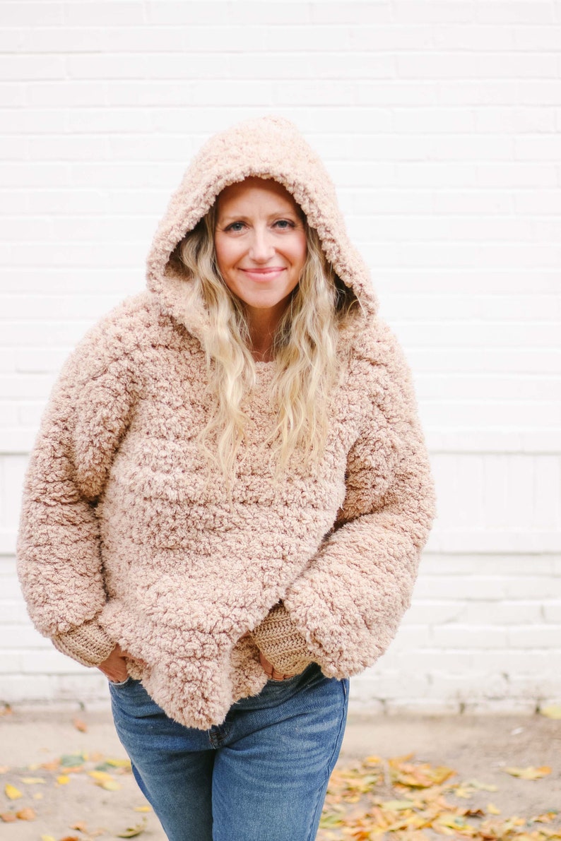 A blonde woman wearing a sherpa hoodie with the hood on her head, and denim jeans with her hands on the front pockets of her jeans.