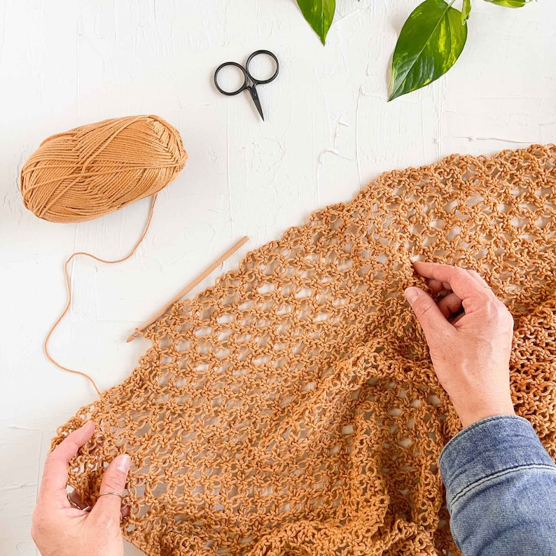 A woman is working on a cotton crochet bag using Solomon's knot stitch, a crochet hook, scissors, and a yarn skein.