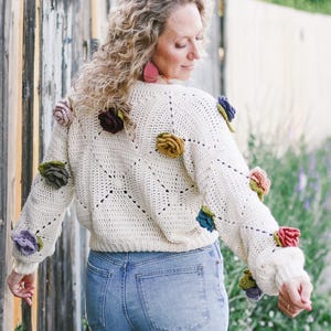 May include: Cream-colored, crocheted sweater adorned with colorful, three-dimensional floral embellishments. The sweater features a square pattern design and is worn with blue jeans. The flowers are in shades of pink, purple, green, and yellow.