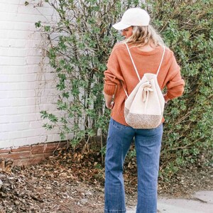 A blonde woman stands with her back to the camera, wearing a baseball cap, an orange sweater, denim jeans, a pair of flats, and a mini backpack on her shoulders.