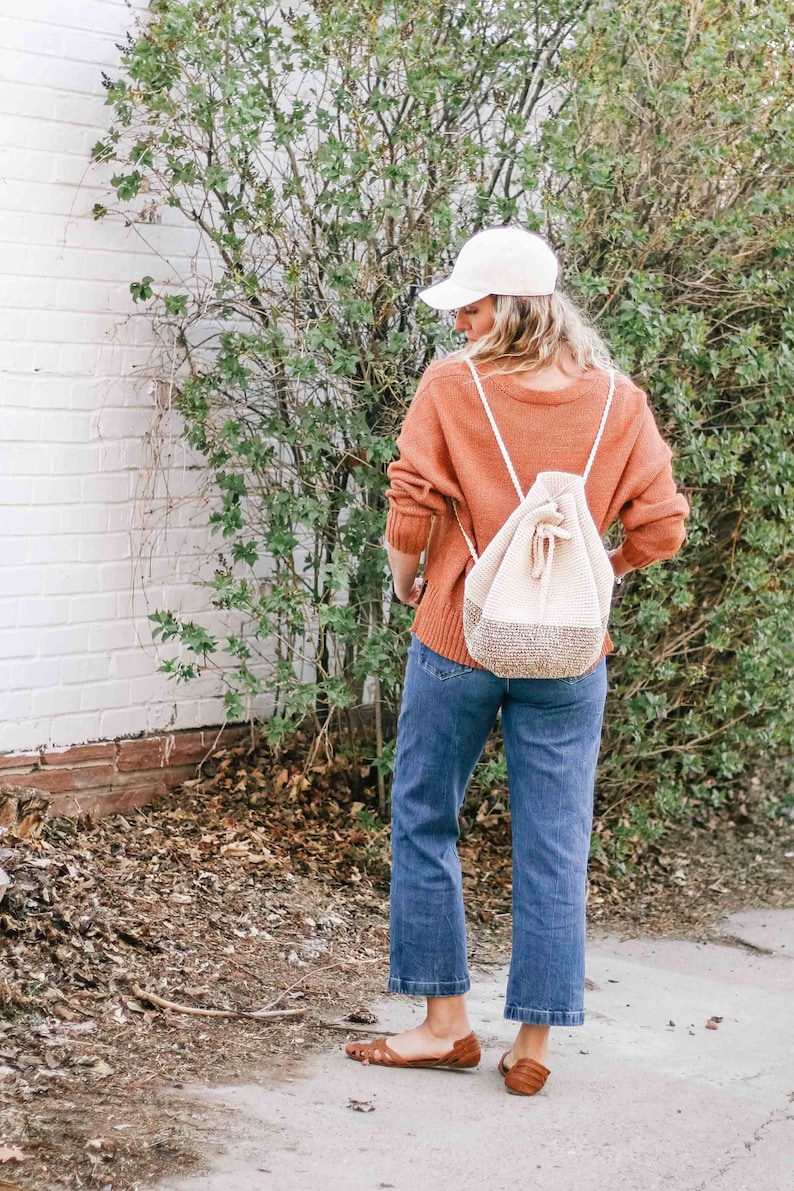 A blonde woman stands with her back to the camera, wearing a baseball cap, an orange sweater, denim jeans, a pair of flats, and a mini backpack on her shoulders.