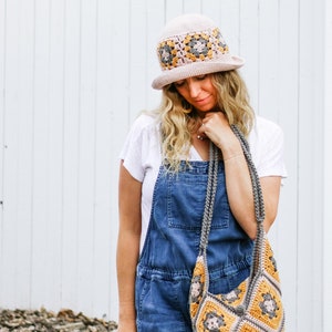 A blonde woman is standing in front of a white wall, looking down while wearing a bucket hat with a matching bag, a white t-shirt, and a denim jumper.