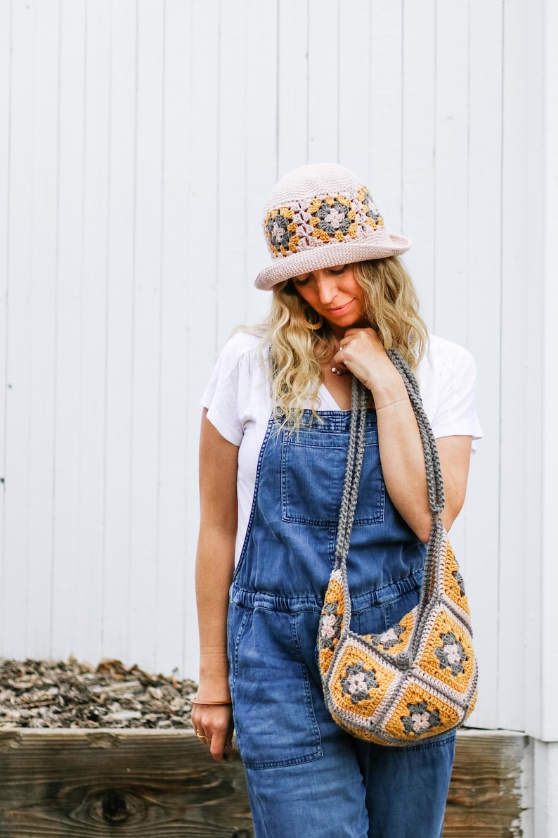 A blonde woman is standing in front of a white wall, looking down while wearing a bucket hat with a matching bag, a white t-shirt, and a denim jumper.