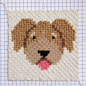 May include: A brown crocheted dog with a pink tongue, stitched on a white gridded background.